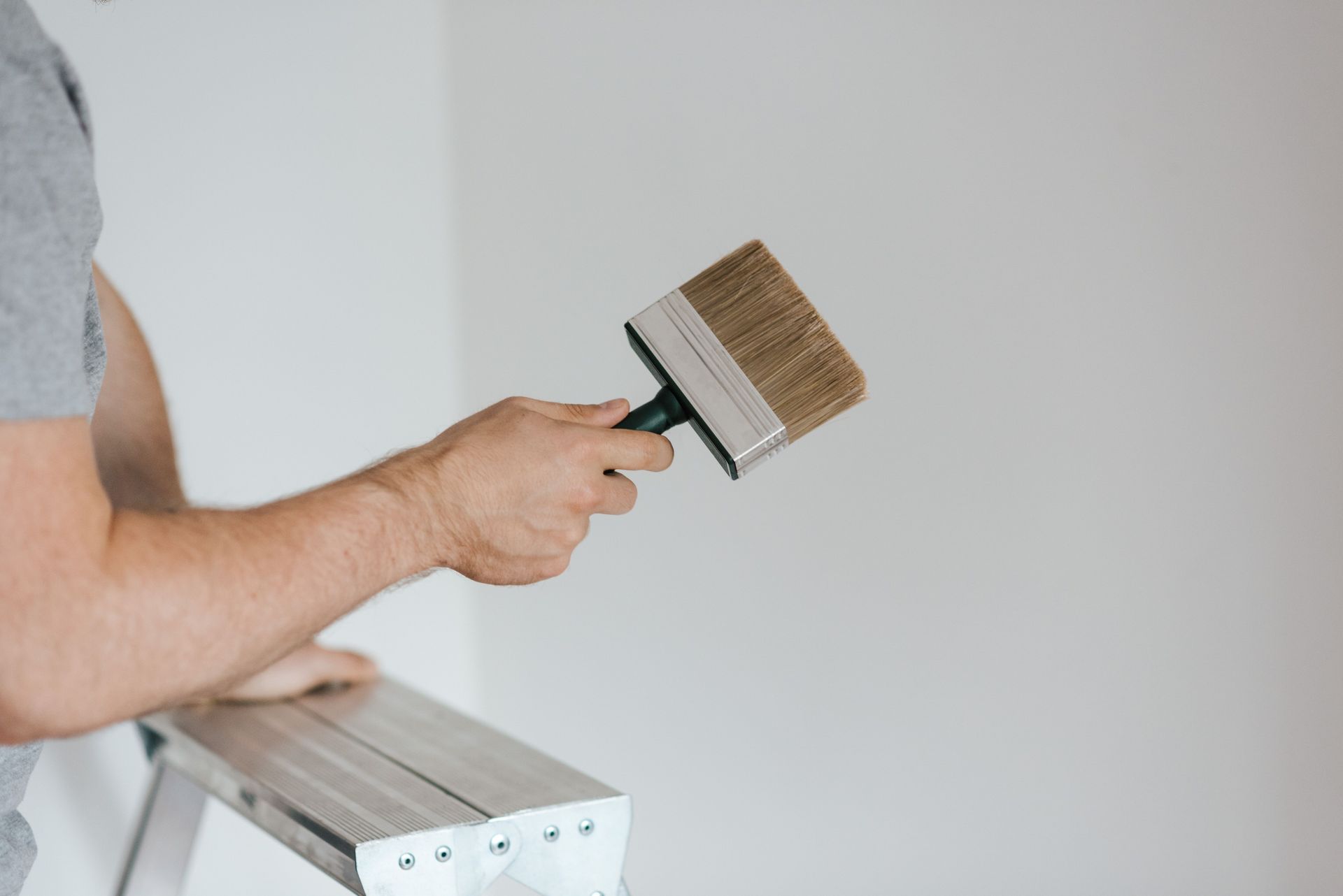 Side view of crop faceless painter in casual clothes holding paintbrush while leaning on stepladder on gray background in soft daylight